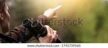 Woman explorer with binoculars showing with finger for animal in the wild forest. Blur dark background. The concept of hunting, travel and outdoor recreation. 