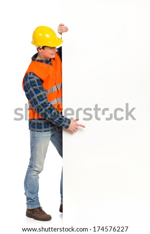 Smiling construction worker in yellow helmet and orange waistcoat standing and pointing at big banner.  Full length studio shot isolated on white.