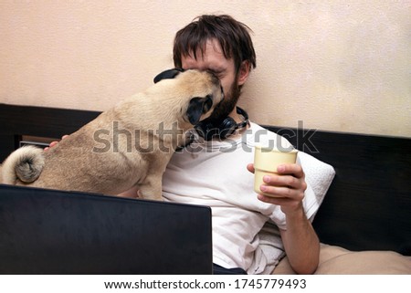 man lying on bed looking at laptop, his pug dog interfere, licking his face