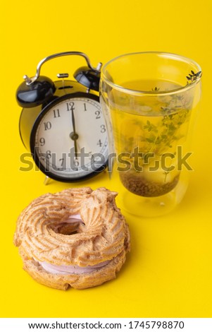 A glass of buckwheat tea with choux ring bun on minimalist bright yellow background, with black analog clock