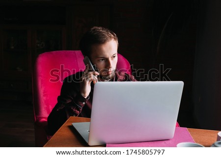 young guy sitting in armchair and talking on the phone on black and white photo. laptop on the table in front of a guy.