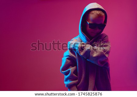 Portrait of a cool boy child in a rap image, stylishly posing in a hoodie, sunglasses and on a neon background.