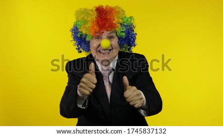 Senior clown director office worker in wig and business suit at work show thumbs up. Businesswoman freelancer boss smiles, looks at camera. Halloween. Yellow background