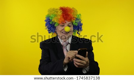 Senior clown office manager worker in wig and business suit using app on smartphone for online distance remote work. Businesswoman freelancer director. Copy space. Halloween. Yellow background