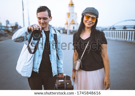 Happy tourists with suitcase on embankment of river. Young man takes photo. Woman wearing casual clothes smiling looking at camera. Traveling concept.