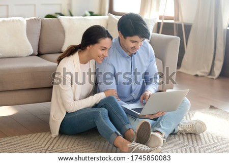 Happy young Caucasian couple relax on floor in living room using modern laptop gadget together, smiling millennial man and woman rest at home browsing wireless internet shopping online on computer