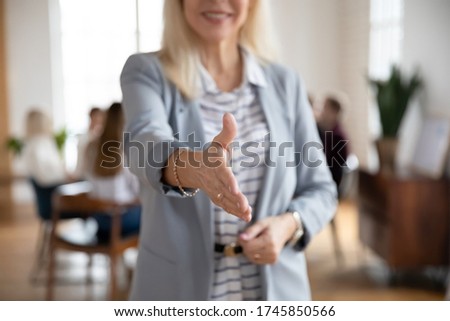 Focus on smiling middle aged blonde businesswoman reaching out hand, greeting partners at office or proposing deal. Happy mature female manager professional welcoming clients, acquaintance concept.