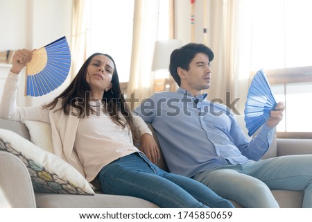 Exhausted young man and woman sit on couch at home breathe fresh air form waver, lack air conditioner, overheated couple rest on sofa suffer from heatstroke or hot weather, wave with hand fan Royalty-Free Stock Photo #1745850659