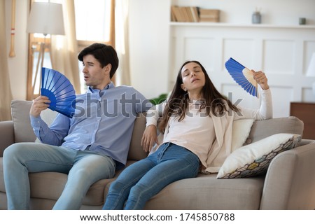Unwell young couple sit relax on couch at home use hand waver suffer from hot weather heatstroke indoors, exhausted overheated man and woman breathe fresh air wave with fan, lack air conditioner Royalty-Free Stock Photo #1745850788