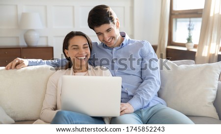 Overjoyed young caucasian couple sit on sofa at home laugh have fun watching funny video online on laptop, happy millennial man and woman relax on couch at home smile using computer together