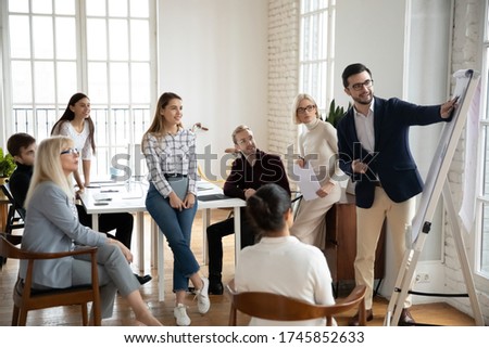 Smiling middle aged and young focused managers listening to motivated male speaker lecturer at workshop. Happy businessman coach trainer showing marketing research results on flipchart to colleagues. Royalty-Free Stock Photo #1745852633