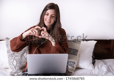 Caucasian young woman making a video call with her family and doing a heart symbol with her fingers. Stay home and social distance to prevent covid-19.