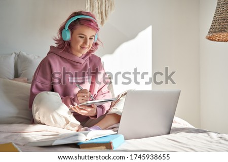 Hipster teen girl school student with pink hair wear headphone write notes watch video online webinar learn on laptop sit in bed distance elearning course video conference pc call in bedroom at home. Royalty-Free Stock Photo #1745938655