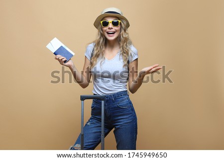 Cheerful blonde woman in sunglasses posing with baggage and holding passport with tickets over beige background.