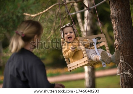 Cute baby girl in bonnet cap is swinging on a wooden swings and her grandmother is playing with her and making funny faces. Image with selective focus and toning