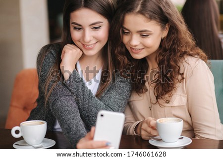 Close up of Two girls friend drink coffee in cafe and look in smat phone. Happy smiled women sittng on terrace. People look stories. Attractive girls