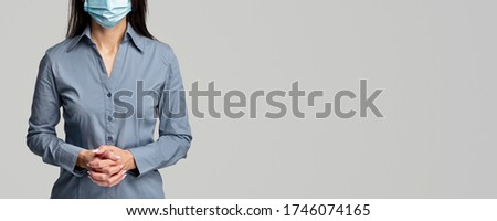 Unrecognizable female in surgical mask clasping hands and standing near blank space on gray background during coronavirus epidemic