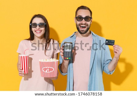 Excited couple friends guy girl in 3d glasses isolated on yellow background. People in cinema concept. Watching movie film, hold popcorn cup soda bank terminal to process acquire credit card payments