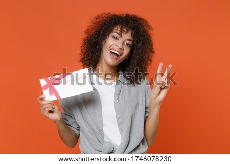 Funny young african american woman girl in gray casual clothes isolated on orange background studio portrait. People lifestyle concept. Mock up copy space. Hold gift certificate, showing victory sign