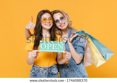 Smiling women girls friends in denim clothes eyeglasses isolated on yellow background. People lifestyle concept. Hold package bag with purchases after shopping, sign with open title showing thumb up