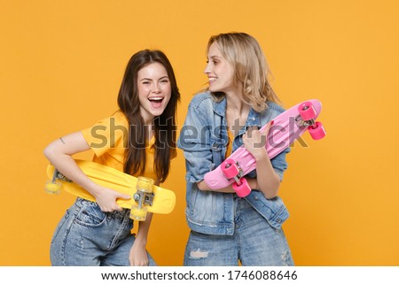 Two laughing young women girls friends in casual t-shirts denim clothes isolated on yellow background studio portrait. People sincere emotions lifestyle concept. Mock up copy space. Hold skateboards