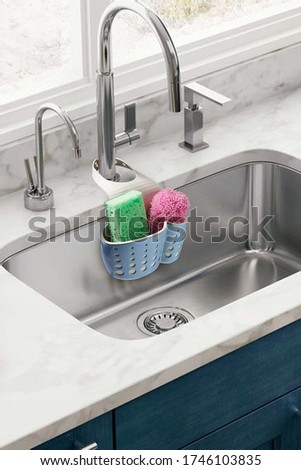 Detailed shot of a blue plastic sponge holder with an adjustable strap. The kitchen organizer with green and pink sponges is hanging on the chrome faucet over the kitchen sink.  Royalty-Free Stock Photo #1746103835
