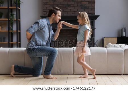 Loving happy young father kneel kiss little princess daughter hand play in living room together, overjoyed dad have fun engaged in funny girly game activity with overjoyed small preschooler girl child