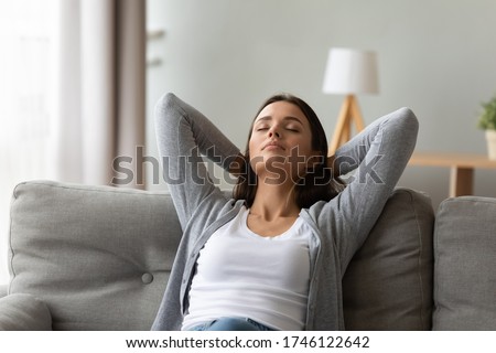Calm 20s woman resting seated on couch in light warm living room put hands behind head enjoy fresh air-conditioned climate control, meditation, inner balance, relaxation at home free lazy day concept Royalty-Free Stock Photo #1746122642