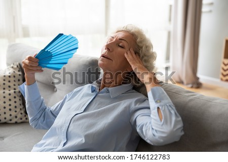 Stressed overheated elderly female leaned on couch holds blue colour fan relieving high temperature refreshing herself in living room without air-conditioner summer weather, hormone imbalance concept Royalty-Free Stock Photo #1746122783
