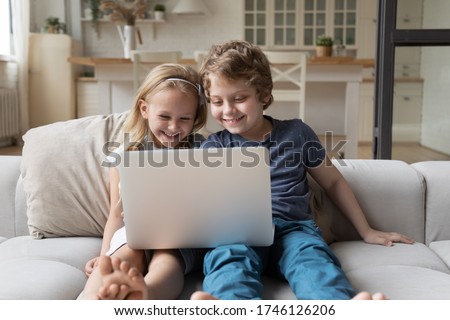 Smiling smart little boy and girl kids sit on couch in kitchen have fun using laptop together, happy small preschooler siblings brother and sister laugh watch funny videos on computer gadget at home
