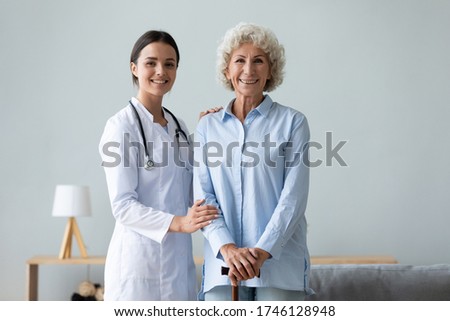 Elderly woman patient holds walking stick photo shooting with young caregiver practitioner in white coat portrait in living room, successful physiotherapy after accident, homecare and nursing concept Royalty-Free Stock Photo #1746128948