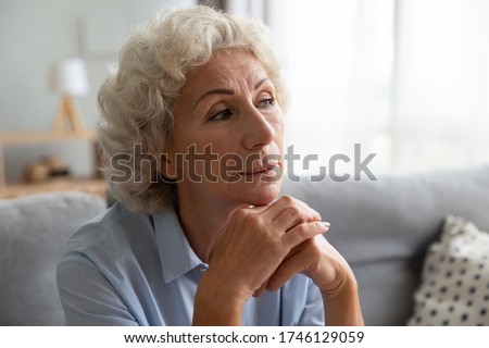 Close up portrait elderly attractive woman seated on couch put chin on folded hands looks pensive lost in nostalgic thoughts, daydreaming, remember recollects life moments, generation of baby boomer Royalty-Free Stock Photo #1746129059