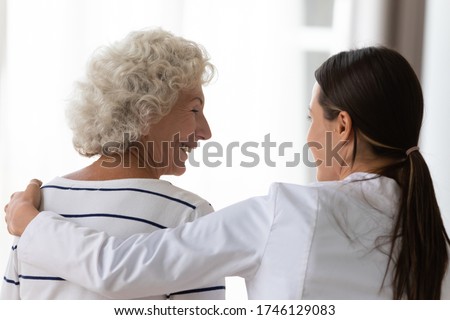 Rear view profile smiling face of satisfied aged woman clinic patient receiving support encouragement from caring young nurse in white coat, caregiver provide help hug old granny during visit at home Royalty-Free Stock Photo #1746129083