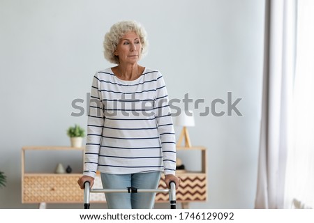 Elderly woman stands in living room alone holds walking frame do exercises, rehab after accident using helpful equipment, senile diseases movement disorder Parkinson Dementia or Osteoporosis, concept Royalty-Free Stock Photo #1746129092