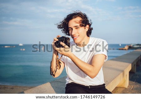 Portrait of carefree teenage tourist holding professional equipment and smiling at camera during hobby time on promenade, happy hipster photographer using technology for making beautiful images