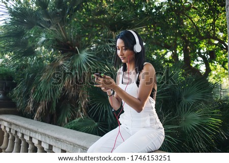Pensive caucasian female millennial using mobile phone for searching and downloading music to playlist on free time outdoors, beautiful woman meloman searching app for enjoying online radio on street