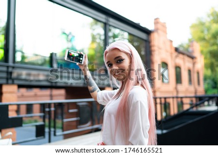 Half length portrait of cheerful millennial blogger with colored pink hair smiling at camera while creating media content via cellular technology, happy influencer with smartphone shooting video