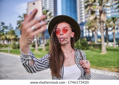 Gorgeous joyful funny modern carefree young hipster woman wearing felt hat and bright red glasses showing tongue during taking selfie photo portrait outdoors 