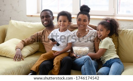 African spouses and little pretty kids spend lazy weekend together seated on couch in living room eating pop-corn choosing show or movie on TV. Happy family activity at home, free time and fun concept