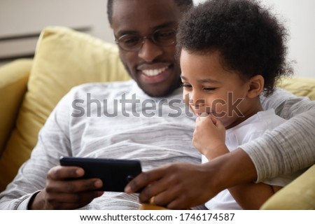 Close up American caring dad spend time with little son family sitting on sofa using smartphone watching cartoons having fun enjoy on-line app for kids. Parental control lazy weekends activity concept