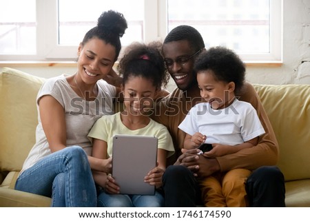 Beautiful full African family spend free time using tablet device watching cartoon educational app for kids enjoy on-line services making videocall chatting with relatives sit on couch in living room
