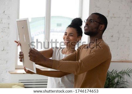African ethnicity millennial couple holding frame admires painting or photo at relocation day at new first home. Concept of remodeling renovation, new furniture store, bank loan, tenancy rented flat