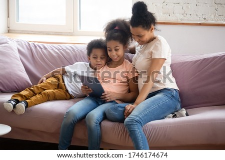 African young mother spend free time at home with pre-teen daughter and little son seated on couch using tablet gadget, children reading using education app website, have fun modern tech usage concept