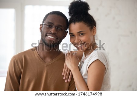 Head shot happy African ethnicity spouses portrait concept. Newly weds pose in rented flat, bank approved loan for young family. Pretty wife snuggles to husband in glasses people smile look at camera