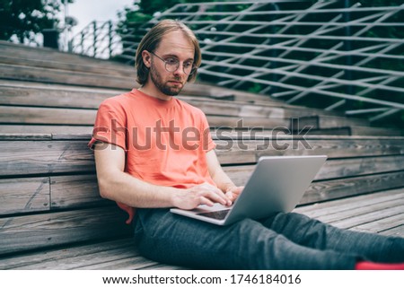 Young man in glasses concentrating on screen of laptop while sitting on wooden steps while working on street in daylight