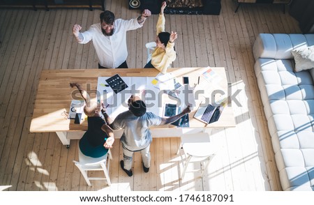 From above trendy modern multiethnic adult employees in casual clothes rejoicing in success while raising arms up at large wooden table