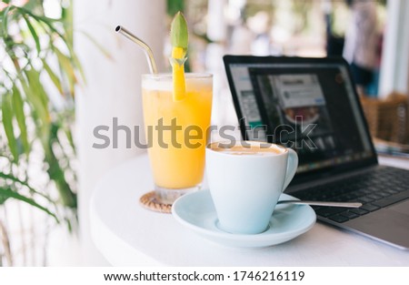 Cropped image of mug with coffee and orange juice for french breakfast near laptop computer on table, close up picture of fresh organic smoothie with straw in glass and caffeine cappuccino cup