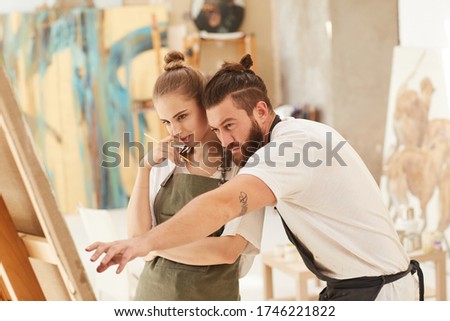 Warm-toned waist up portrait of contemporary couple painting picture together while standing by easel in art studio, copy space