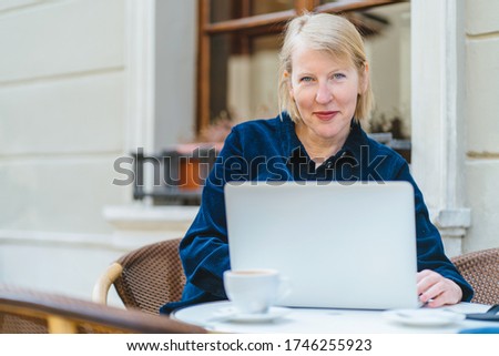Blond mature scandinavian woman in cafe writing on document and using laptop. Your business proposal has just arrived. Smiling senior female open laptop in cafeteria terrace open air.