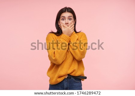 Studio photo of young shocked brunette lady with casual hairstyle covering her mouth with raised palms and looking amazedly at camera with wide eyes opened, posing over pink background